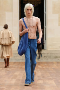 Male model walking runway, shirtless with blue denim jeans, carrying a large blue bag over his shoulder and wearing a beaded necklace.