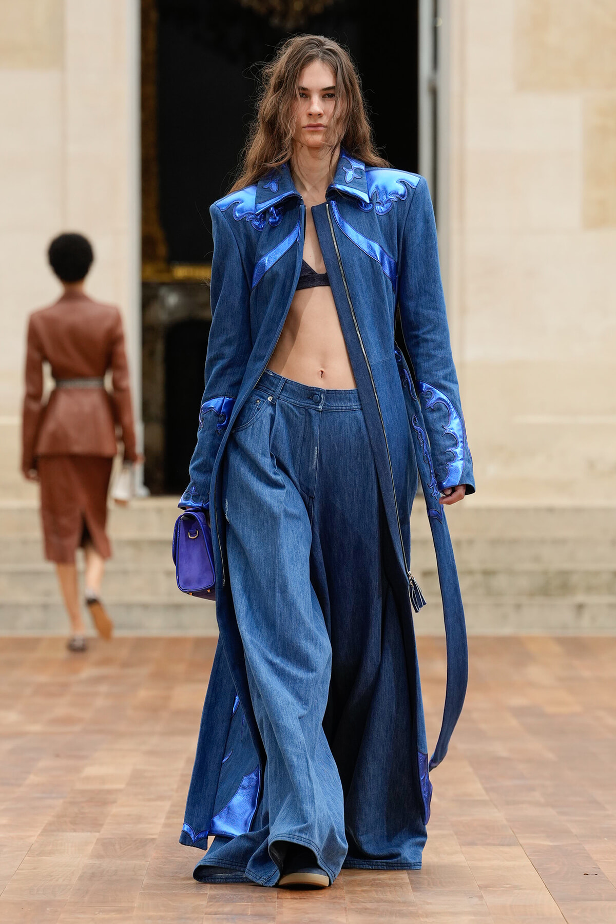 Model walking the runway in an oversized blue denim suit with purple satin trim and a bralette beneath.