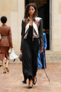 Model on a runway wearing a color-block brown, black, and white leather jacket with matching details, black wide-leg pants, tan ankle boots, and a small brown handbag.