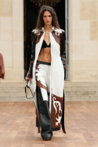 Fashion model walking a runway in a long white coat with brown and black leather panels and decorative white motifs, black bra top underneath, holding a small bag