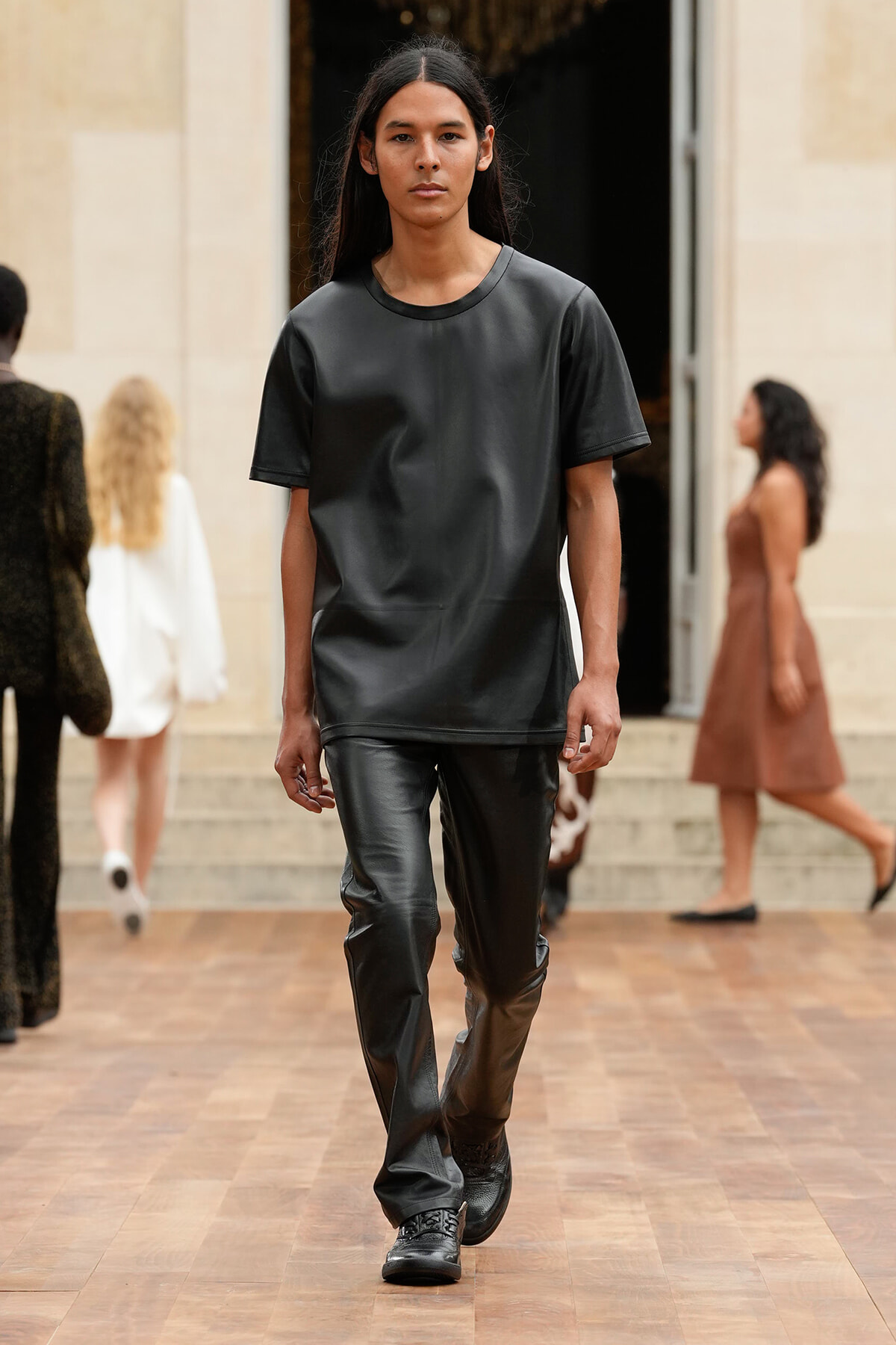 Model walking a fashion runway in an all-black leather-look outfit: loose short-sleeve top and matching pants wearing black shoes.