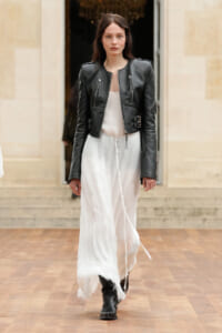 Model wearing a black leather jacket over a flowing white dress, walking on a wooden runway in boots.