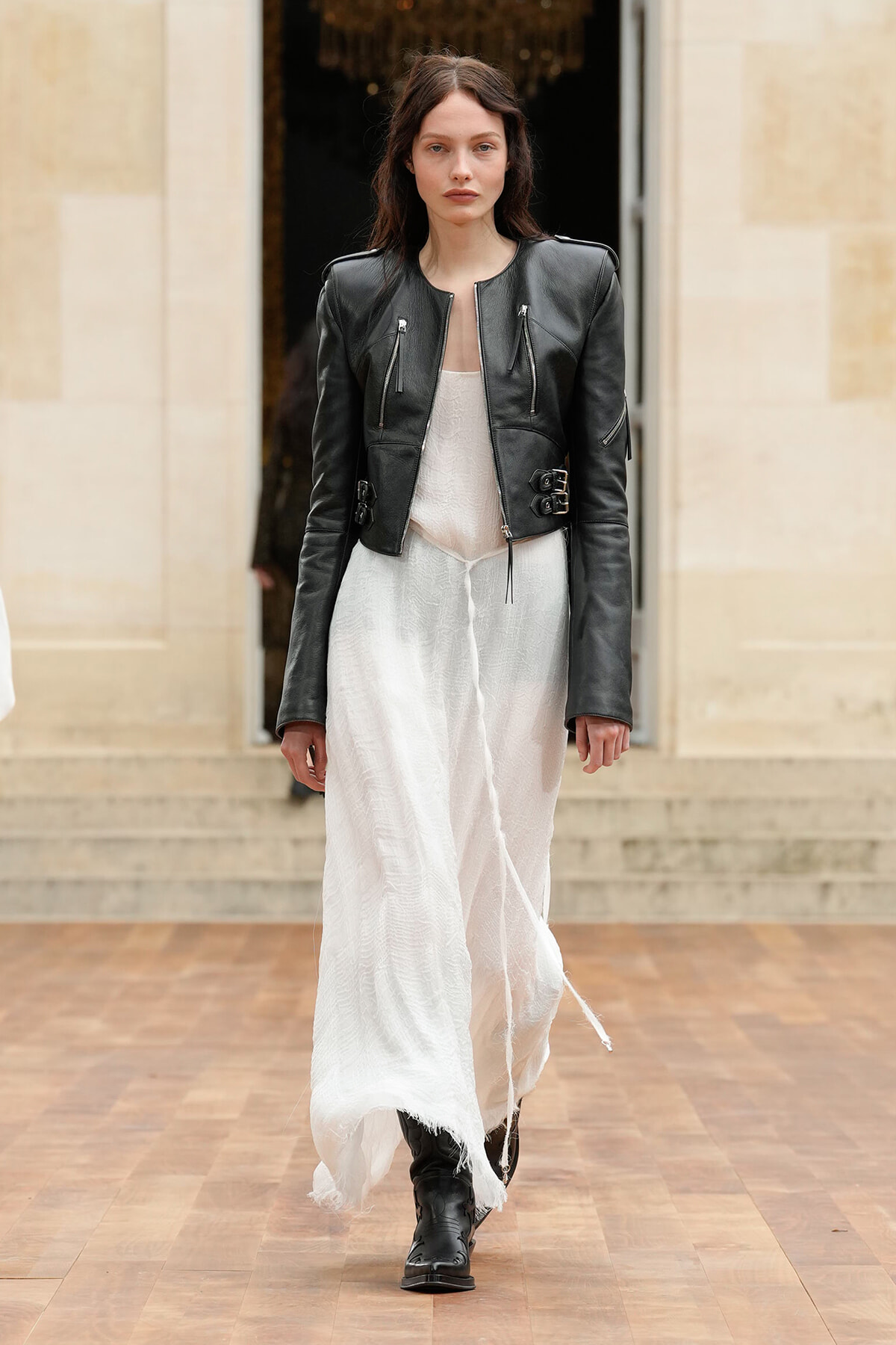 Model wearing a black leather jacket over a flowing white dress, walking on a wooden runway in boots.