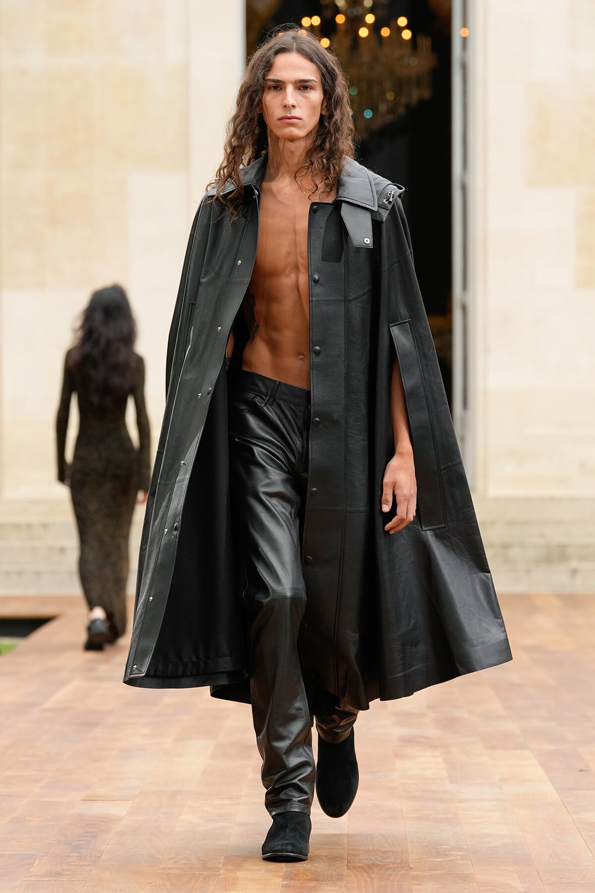 Model walks a runway in an oversized black coat open at the front, revealing a bare chest and black leather pants, with black ankle boots.