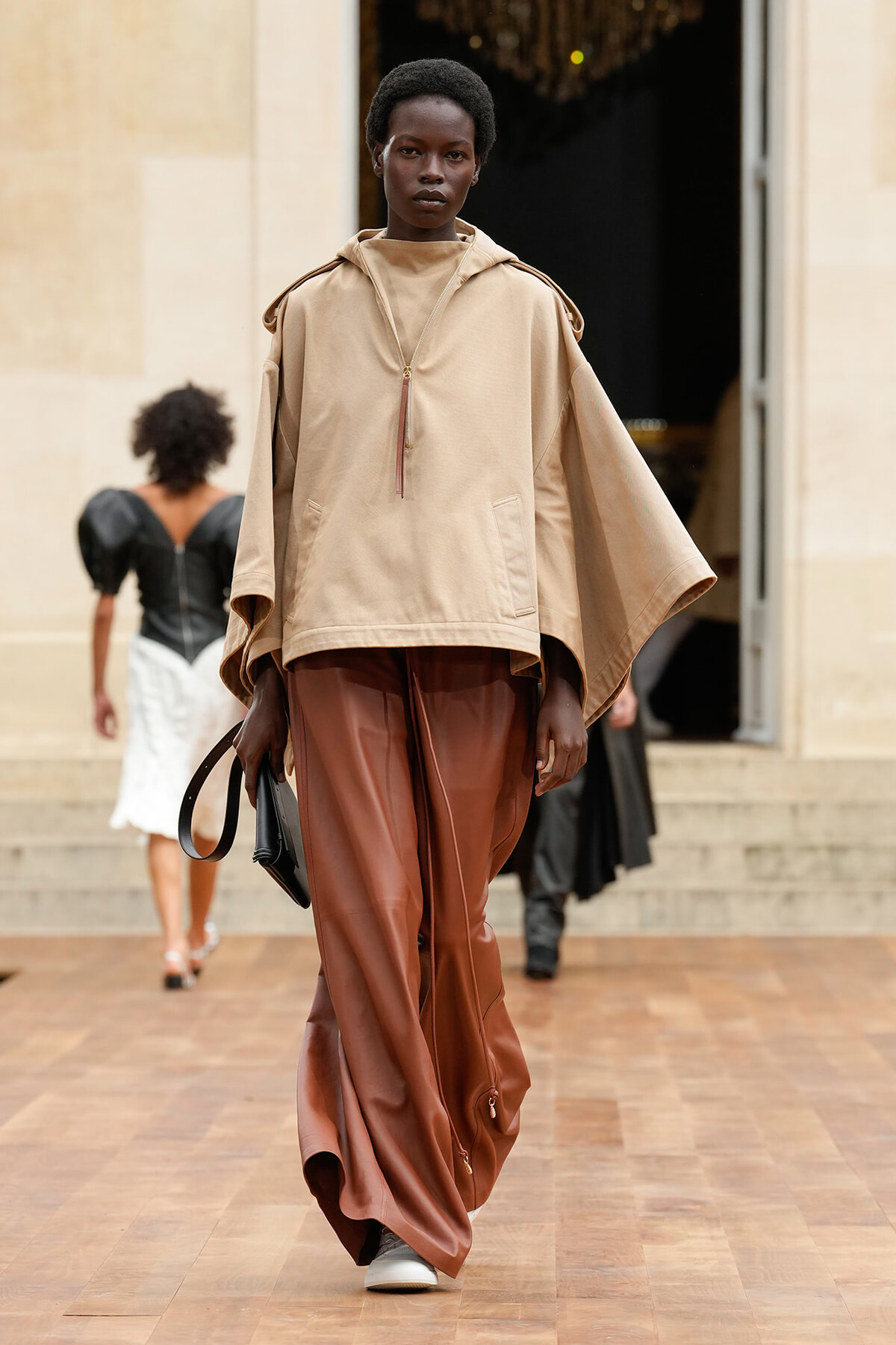 Model walks a fashion runway in a beige cape-style top and rust-brown wide-leg pants, carrying a black handbag.