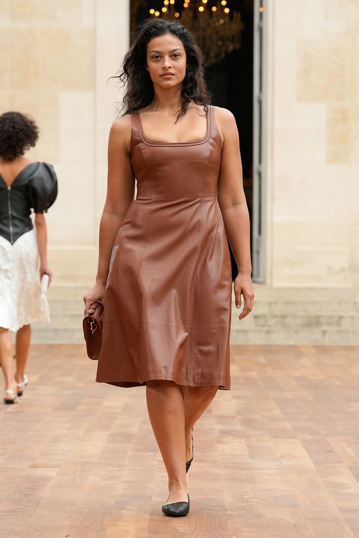 Woman in a brown leather knee-length dress walks toward the camera, carrying a matching brown clutch under her arm.