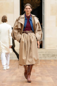 Model walking in a beige trench coat layered over a blue shirt and burgundy top, belted at the waist with brown boots in a fashion show setting.
