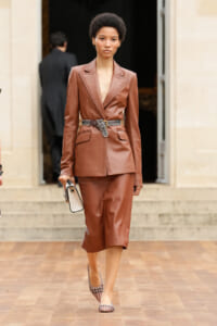 Model in a brown leather blazer and matching midi skirt with a patterned belt, holding a small beige bag as she steps onto a wooden floor outside a building.