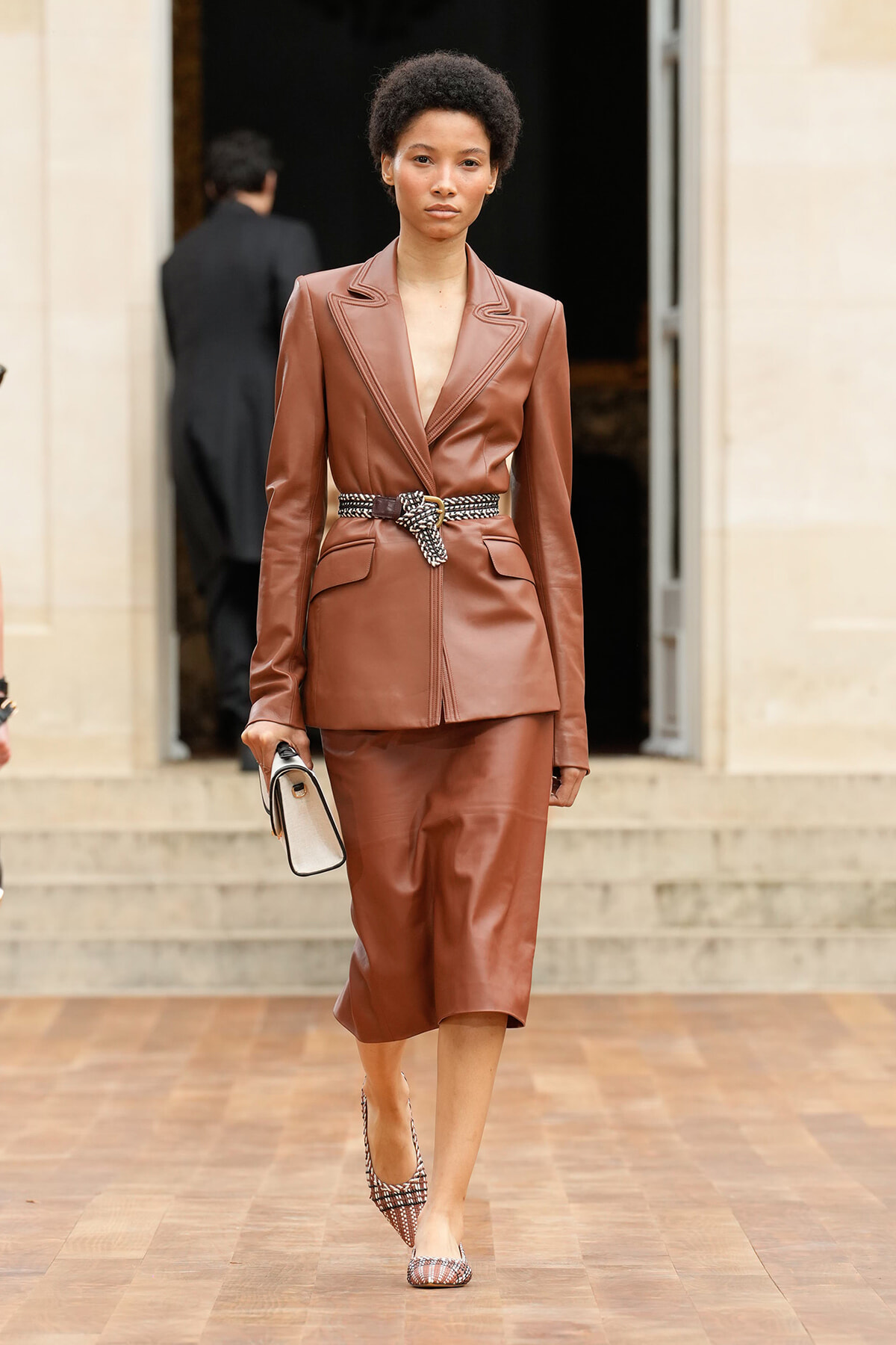 Model in a brown leather blazer and matching midi skirt with a patterned belt, holding a small beige bag as she steps onto a wooden floor outside a building.