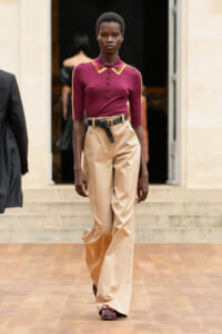 Model walking a runway in a burgundy polo top with tan wide-leg trousers and a black belt