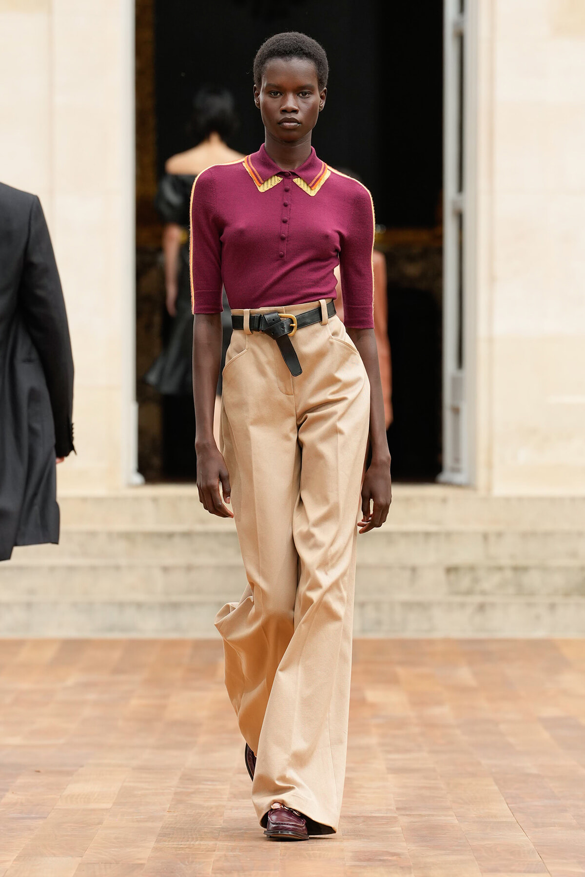 Model walking a runway in a burgundy polo top with tan wide-leg trousers and a black belt