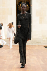 Model walking on a wooden runway in a black glittery jumpsuit with a pearl necklace, outside a stone building.