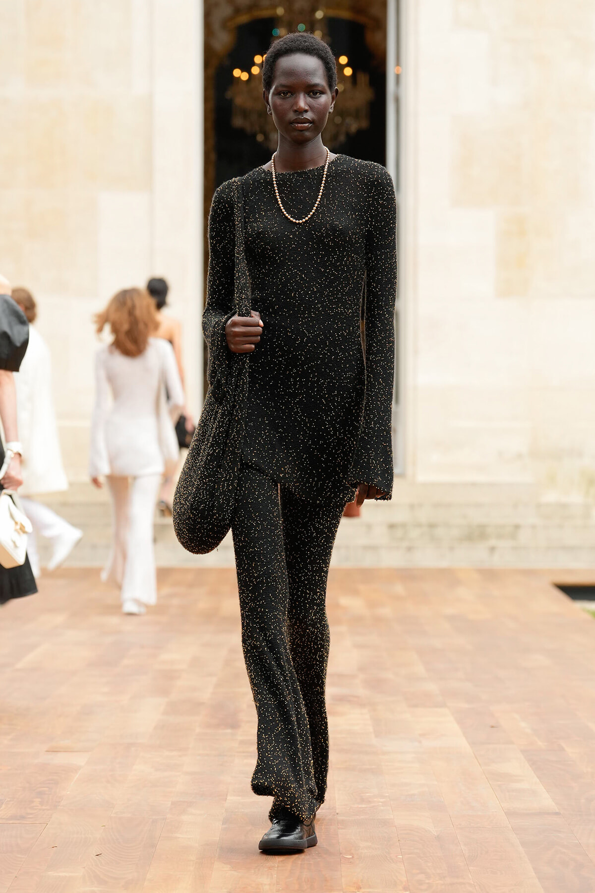 Model walking on a wooden runway in a black glittery jumpsuit with a pearl necklace, outside a stone building.