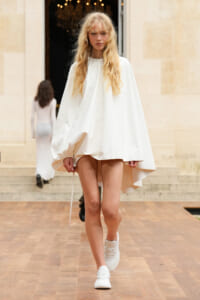 Model walking a runway in a loose white cape dress with blonde hair and white sneakers, set against a stone building backdrop.