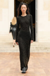 Model walking a fashion runway in a full-length black glittery gown with long sleeves.