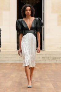 Model walking on a runway in a black V-neck top with voluminous puff sleeves and a white midi skirt, holding a small clutch.