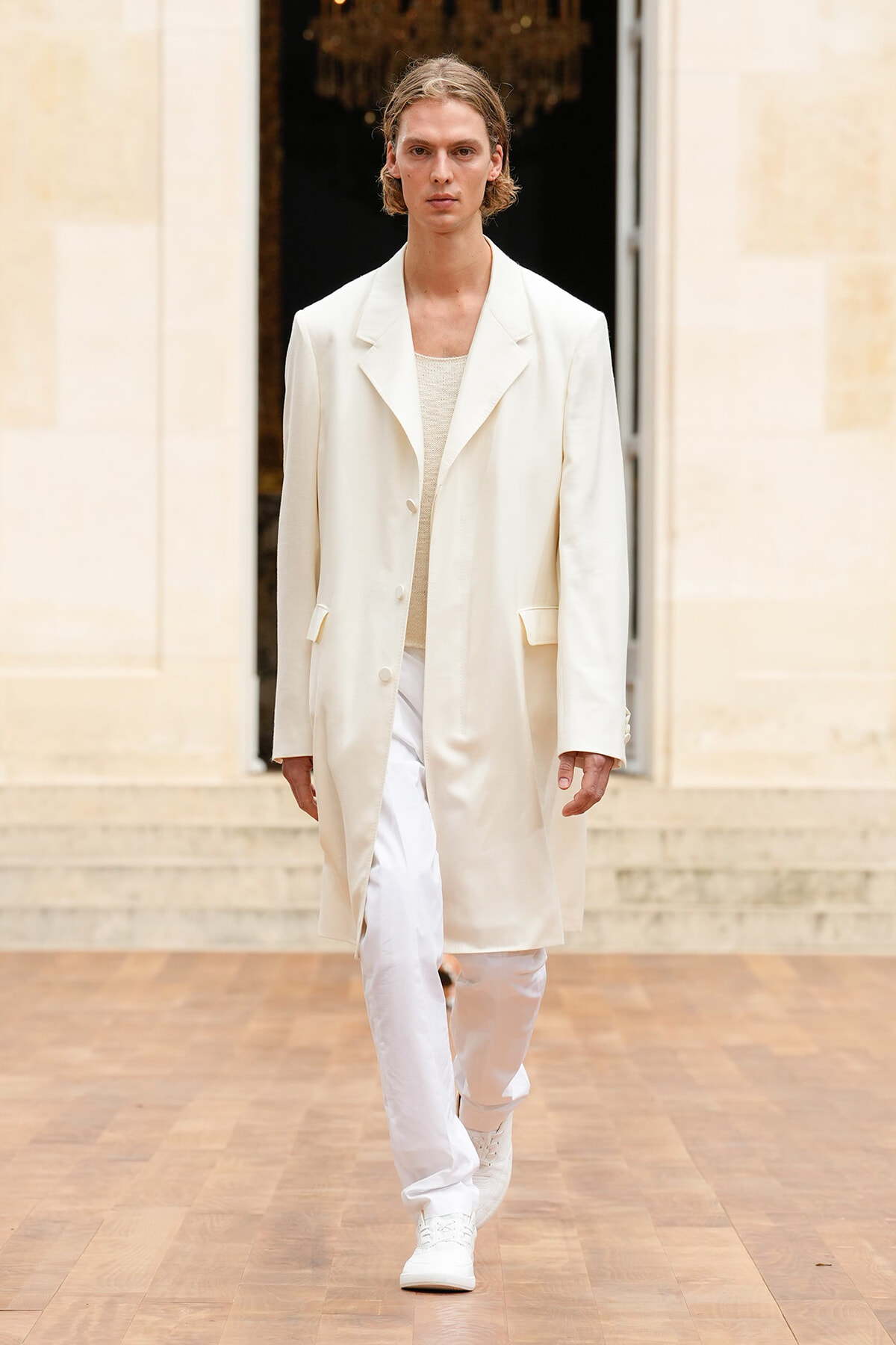 Male model walking a runway in a long cream coat over a beige knit top and white pants, neutral expression.