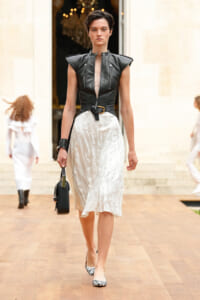 Model walking runway in a black sleeveless leather top with zippers and a light midi skirt, carrying a small black handbag.