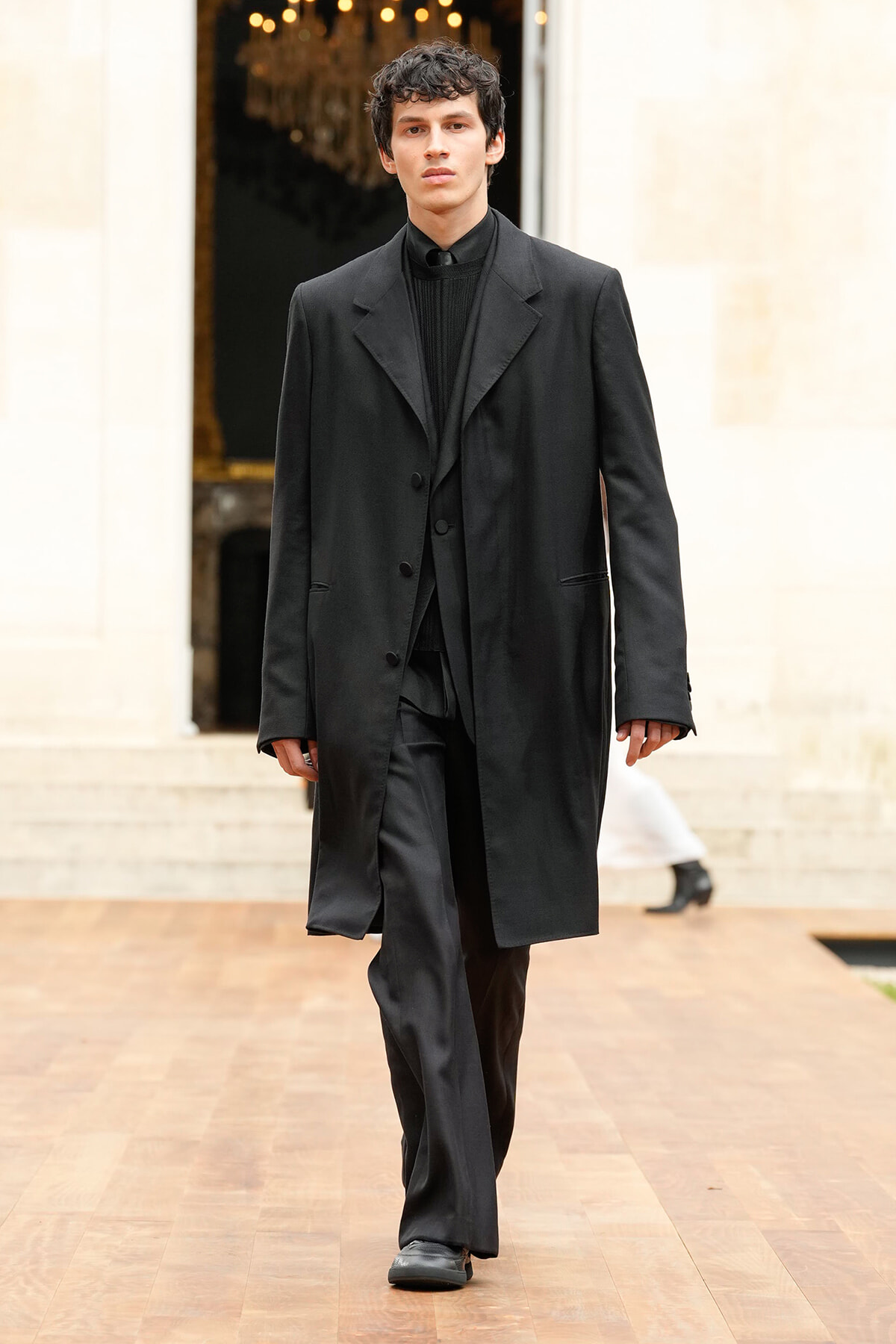 Male model walks a runway in a black overcoat and matching suit, neutral backdrop behind him.