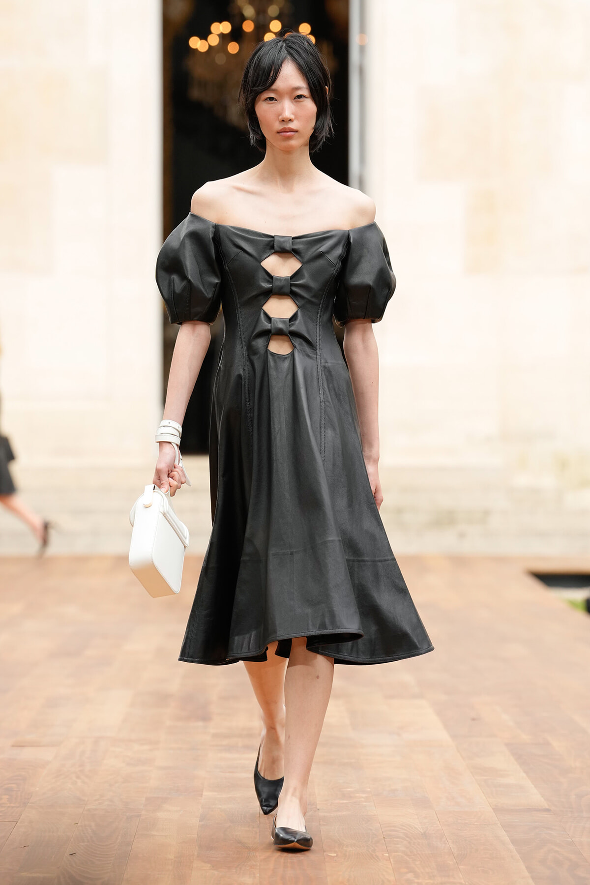 Model wearing a black off-the-shoulder dress with cutouts and puff sleeves, walking on a wooden runway, holding a white handbag.