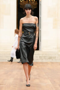 Model walking a fashion runway in a strapless black leather midi dress with a braided belt and a black clutch, showing tattoos on both arms, neutral expression.