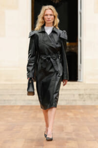 Woman walking forward in a black leather trench coat, belted at the waist, carrying a small handbag and wearing pointed heels.