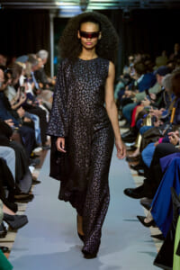 Model walks a fashion show wearing a black leopard-print jumpsuit with a flowing one-shoulder cape and futuristic red sunglasses.