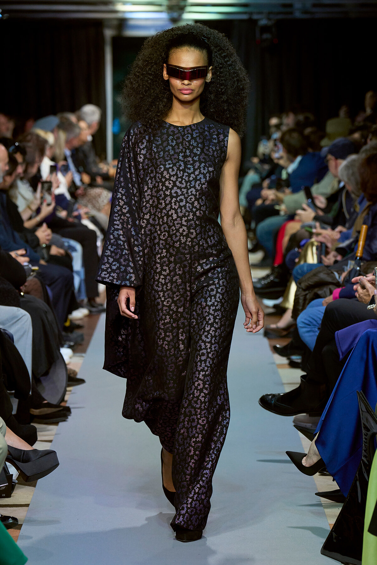 Model walks a fashion show wearing a black leopard-print jumpsuit with a flowing one-shoulder cape and futuristic red sunglasses.