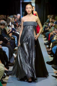 Model walks the runway in a strapless, metallic silver gown with a wide ruched bodice and a flowing, textured skirt; audience watches from sides.