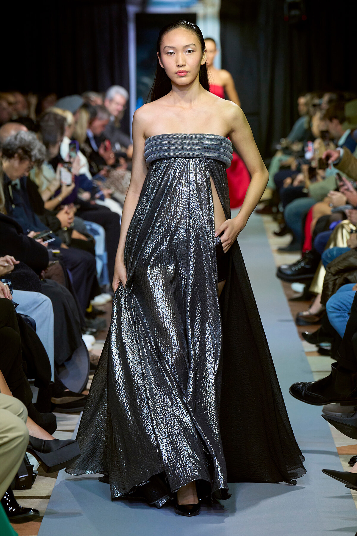 Model walks the runway in a strapless, metallic silver gown with a wide ruched bodice and a flowing, textured skirt; audience watches from sides.
