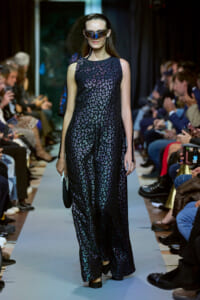 Model on a runway wearing a sleeveless, metallic leopard‑print maxi dress with futuristic visor sunglasses, holding a small clutch.