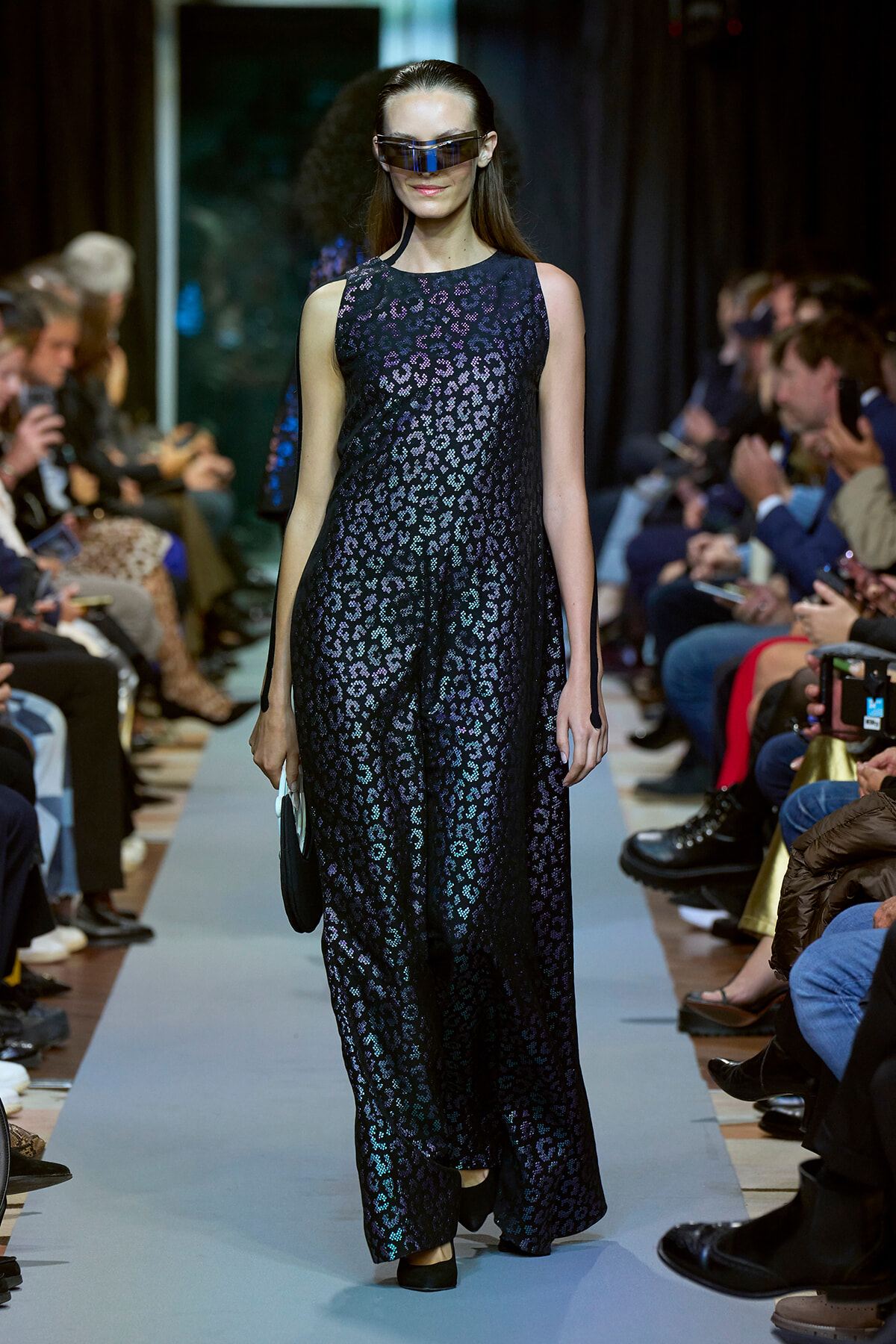 Model on a runway wearing a sleeveless, metallic leopard‑print maxi dress with futuristic visor sunglasses, holding a small clutch.