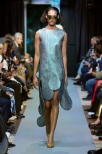 Model walks the runway in a sleeveless metallic blue dress with large circular petal-like panels and gold heels, wearing futuristic sunglasses.