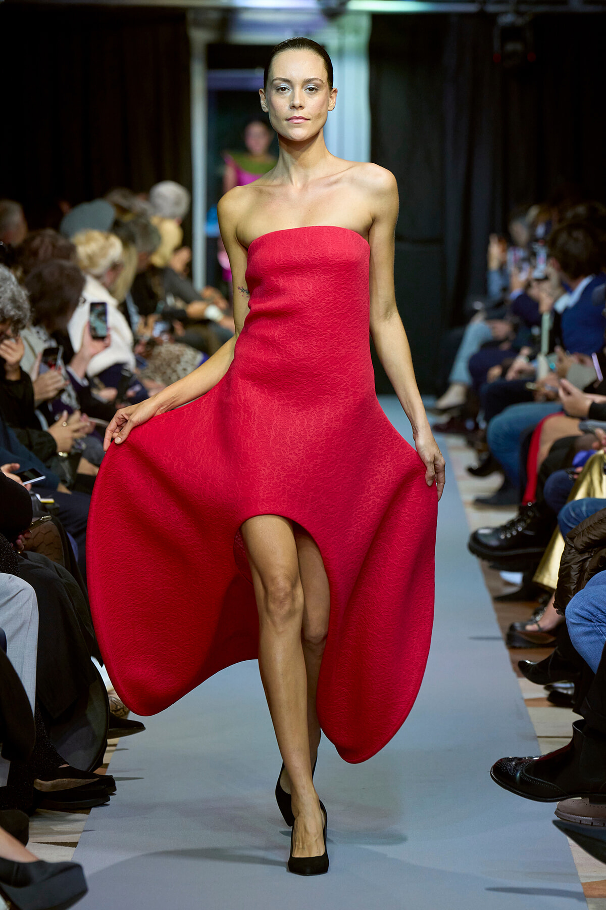 Model walks a bright red strapless dress down a runway, audience on both sides watching.