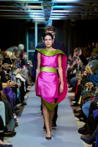 Model walking a fashion runway in a hot-pink satin dress with lime-green accents; audience on both sides filming with phones.