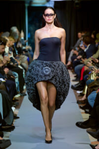 Model walks a fashion runway in a strapless navy dress with a voluminous lace bubble skirt and dark sunglasses, audience in background.