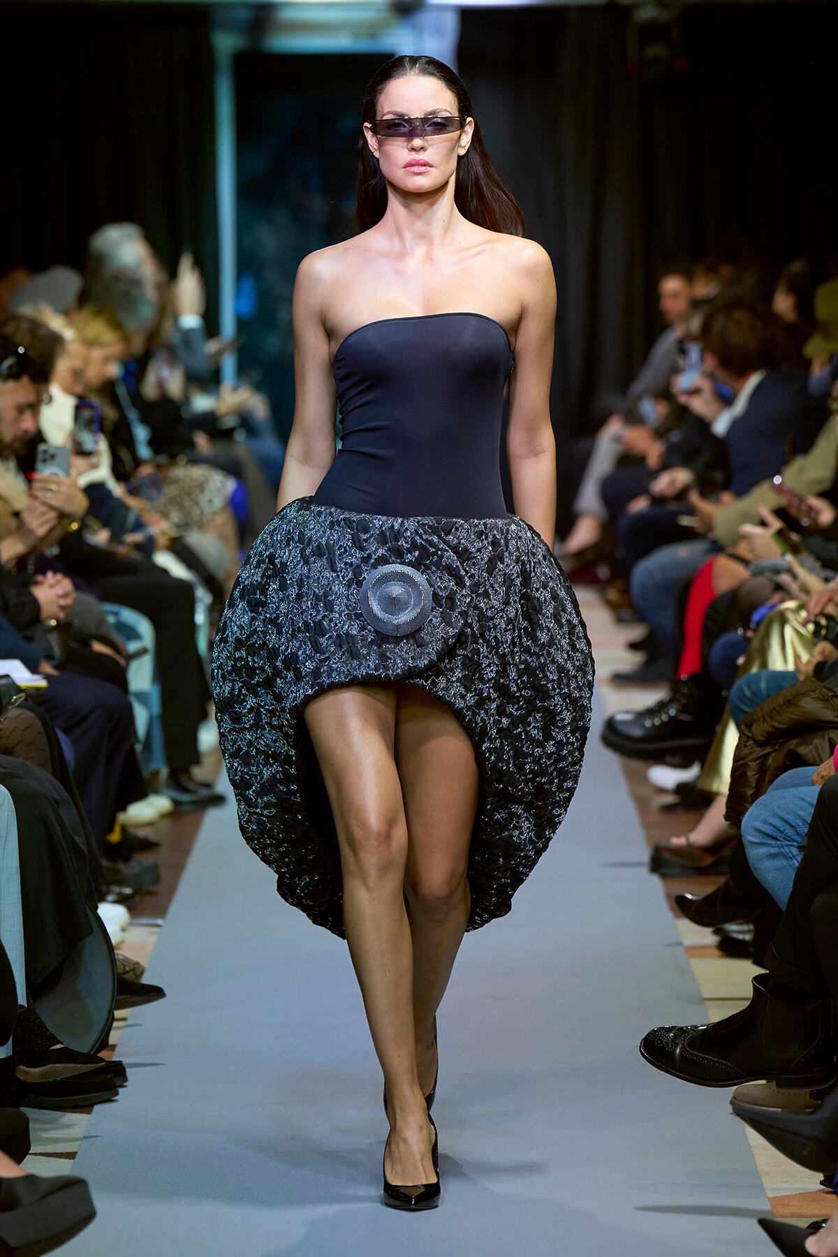 Model walks a fashion runway in a strapless navy dress with a voluminous lace bubble skirt and dark sunglasses, audience in background.