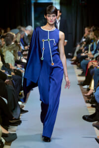 Model walking the runway in a royal blue asymmetrical jumpsuit with white piping and multicolored circular embellishments, audience visible on both sides.