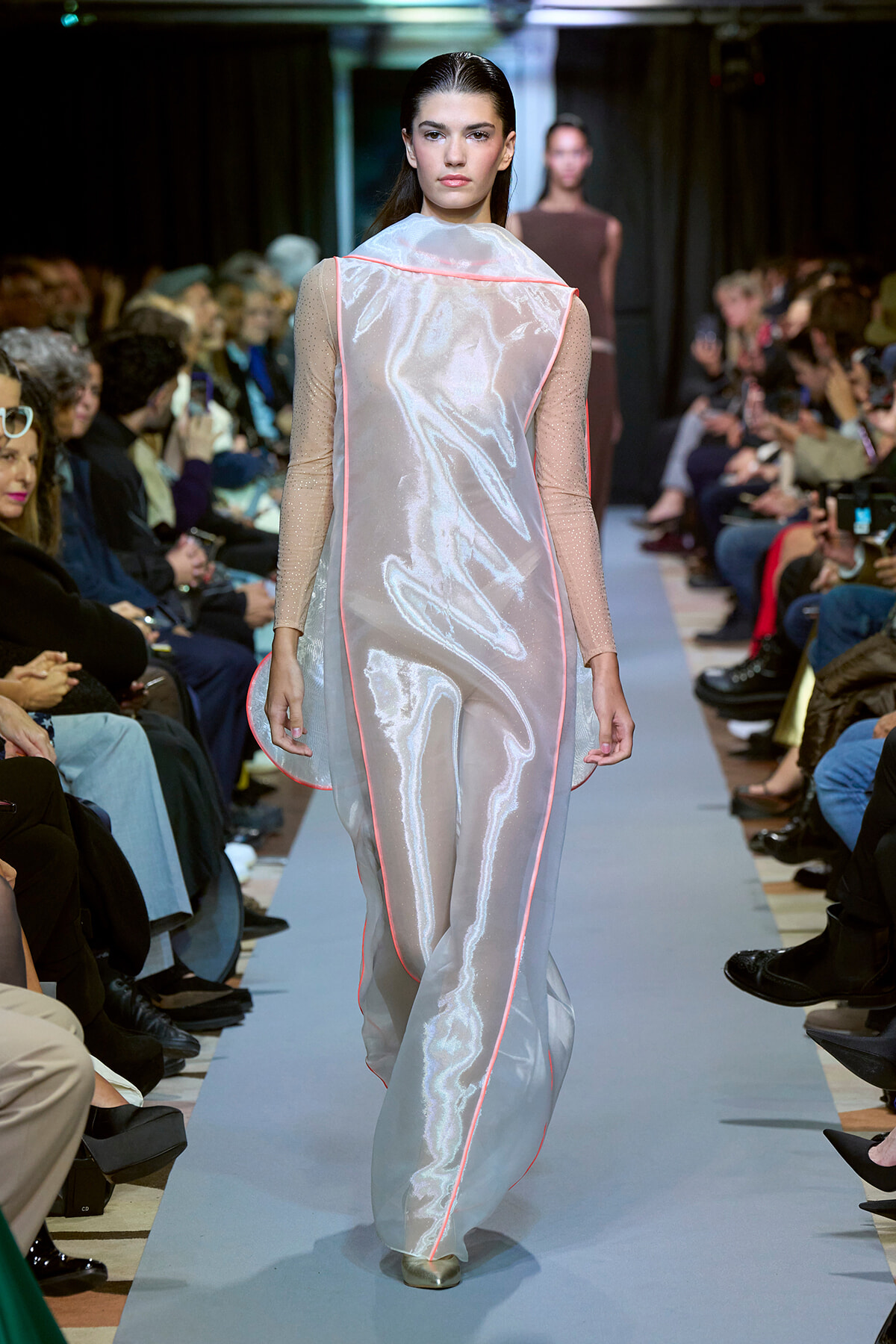 Model walks a fashion runway in a shiny silver gown with pink piping along the seams and sheer sleeves.