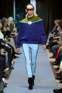 Model on a fashion runway wearing a navy purple zip-up jacket with teal trim and green yoke, light blue pants, and black boots.