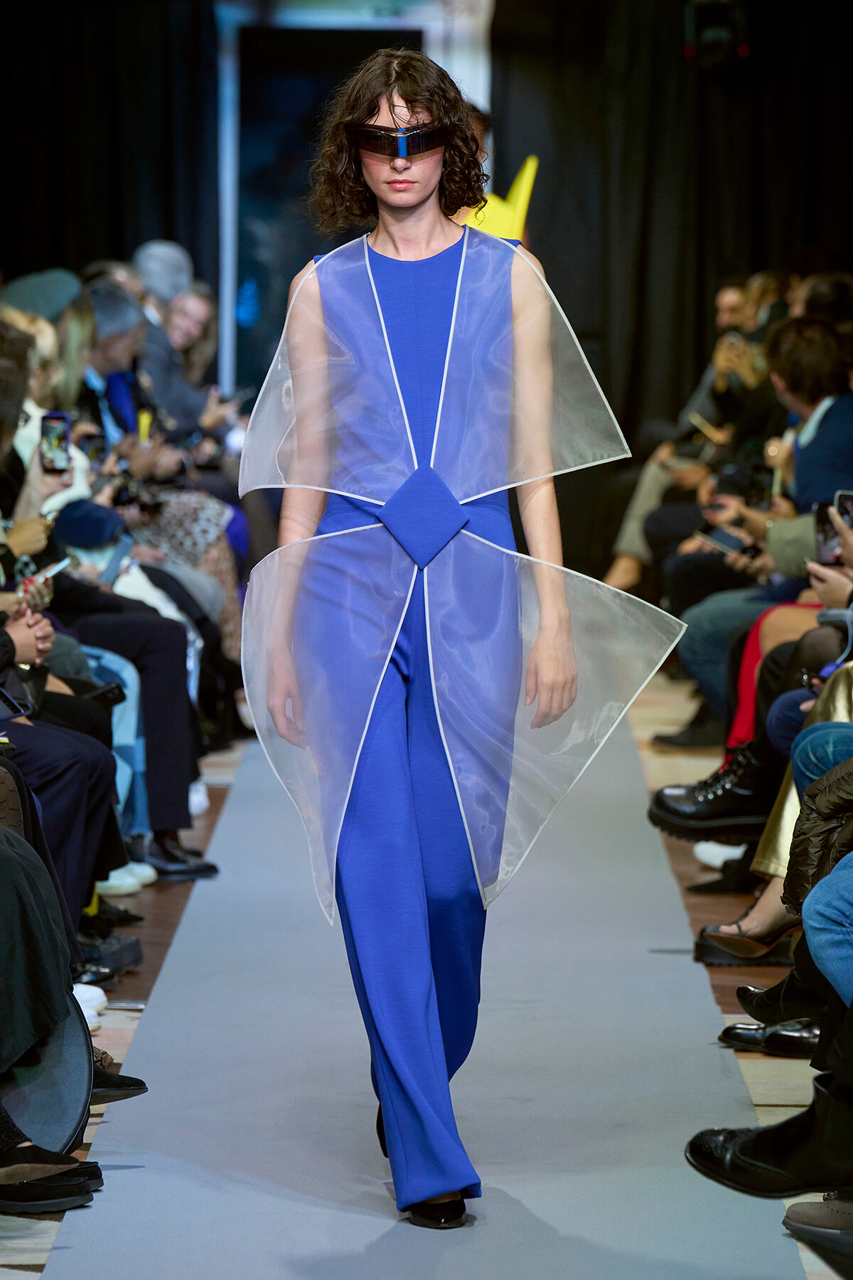 Model walks a fashion show in a blue avant-garde gown with transparent cape panels and a futuristic visor, audience on both sides.