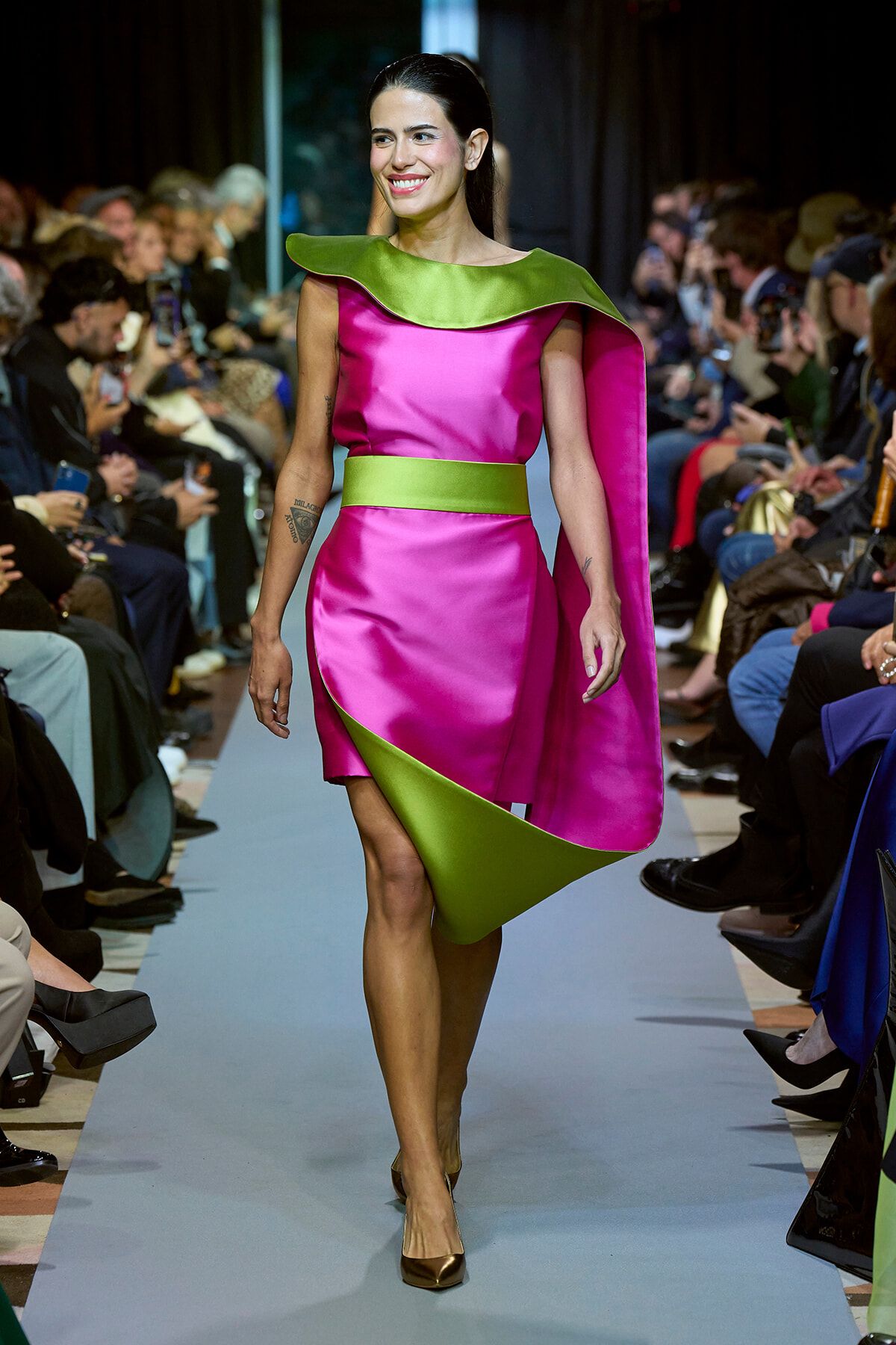 Model walks the runway wearing a hot pink satin dress with a lime green cape, belt, and hem accents, paired with bronze heels, as a fashion show audience watches.