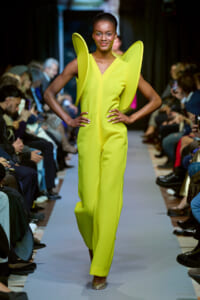 Model walks the runway in a neon yellow jumpsuit with oversized wing-like shoulders and a front zipper.