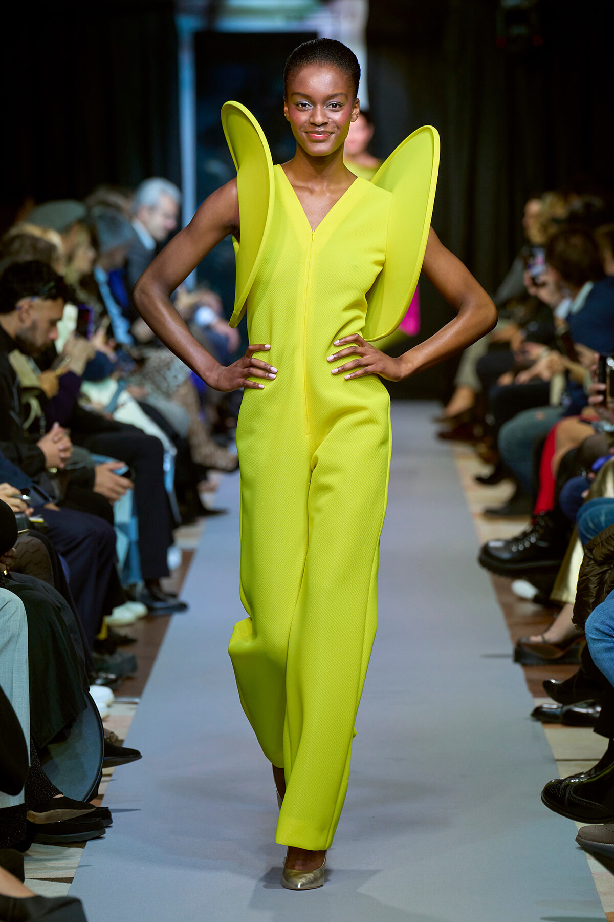 Model walks the runway in a neon yellow jumpsuit with oversized wing-like shoulders and a front zipper.
