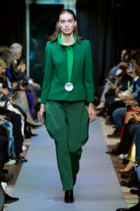 Model on a runway wearing a green tailored suit with a bright green inner panel and a large silver circular accessory at the waist.