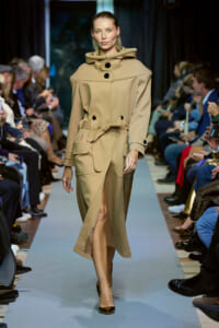 Model walks the runway in a beige oversized trench coat with a large collar, belt at the waist, and brown high heels.
