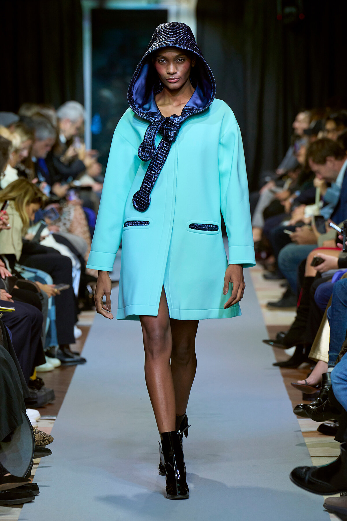 Model walks the runway wearing a turquoise hooded coat with a navy braided scarf and black patent boots in a fashion show.