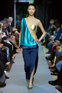 Model walks the runway in a teal satin one-shoulder top with beige mesh and navy pants, wearing a gold circular accessory on her chest.