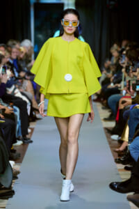 Model walks a runway in a lime-yellow cape-sleeved mini dress with a large white circular accessory at the waist and white ankle boots with yellow sunglasses.