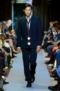 Male model walks the runway wearing a navy suit with a burgundy necktie featuring two white diamond shapes, audience watching on both sides.